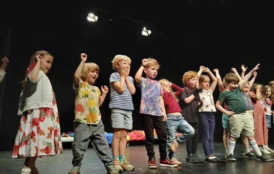 A line of small children at the front of the stage. They are cheering and pretending to watch a race.