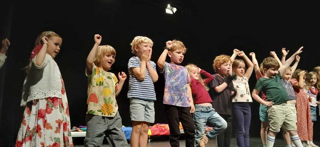A line of small children at the front of the stage. They are cheering and pretending to watch a race.
