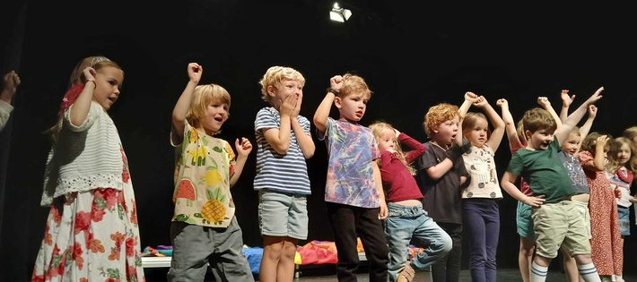 A line of small children at the front of the stage. They are cheering and pretending to watch a race.