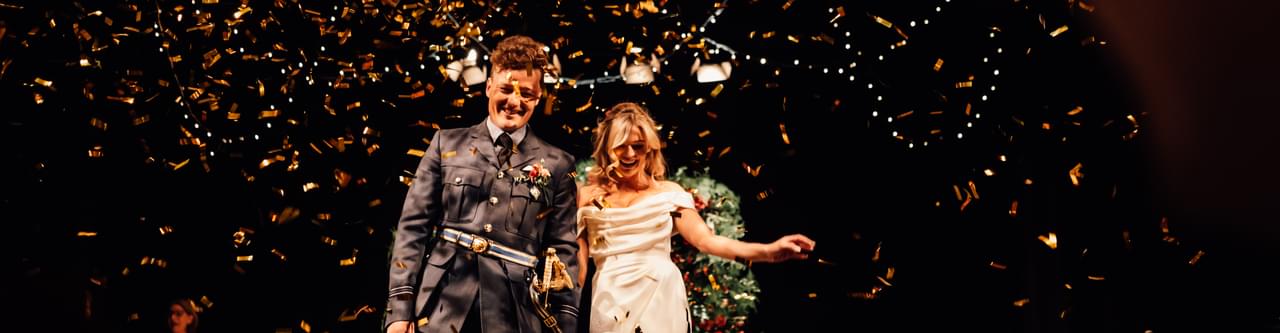 Groom in military uniform and bride in white gown on the Theatre stage as gold confetti rains down around them