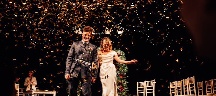 Groom in military uniform and bride in white gown on the Theatre stage as gold confetti rains down around them