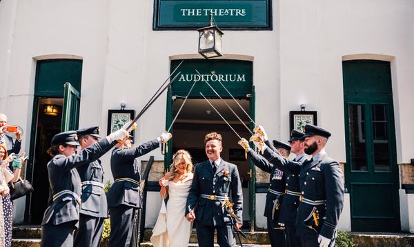 Bride and groom walking through guard of honour of swords on steps outside white Theatre building