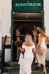 Bride in off the shoulder white gown and bridesmaids in gold enter the auditorium door