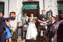 Groom in a kilt and bride in a white gown walk down the Theatre steps as guests throw confetti