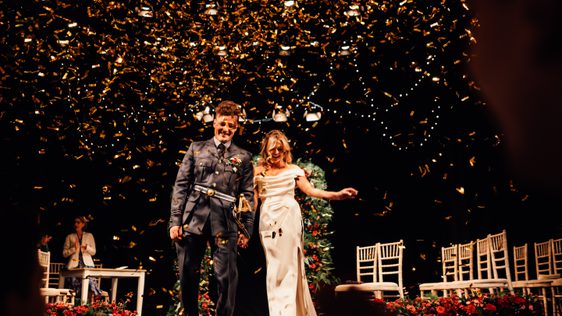 Groom in military uniform and bride in white gown on the Theatre stage as gold confetti rains down around them