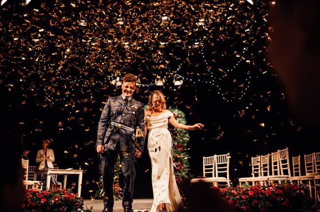 Groom in military uniform and bride in white gown on the Theatre stage as gold confetti rains down around them