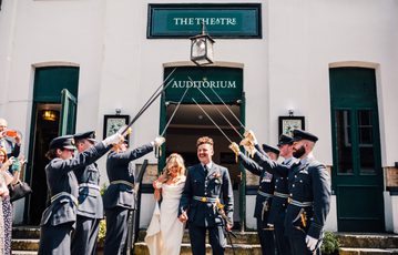 Bride and groom walking through guard of honour of swords on steps outside white Theatre building